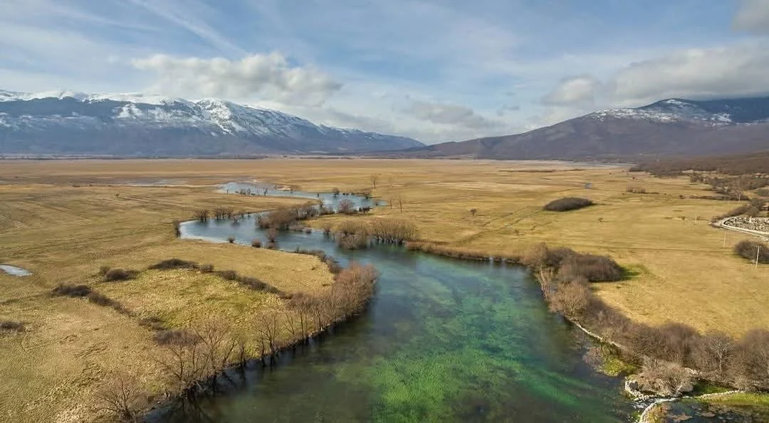 A river winding through the polje, snow-covered mountains behind